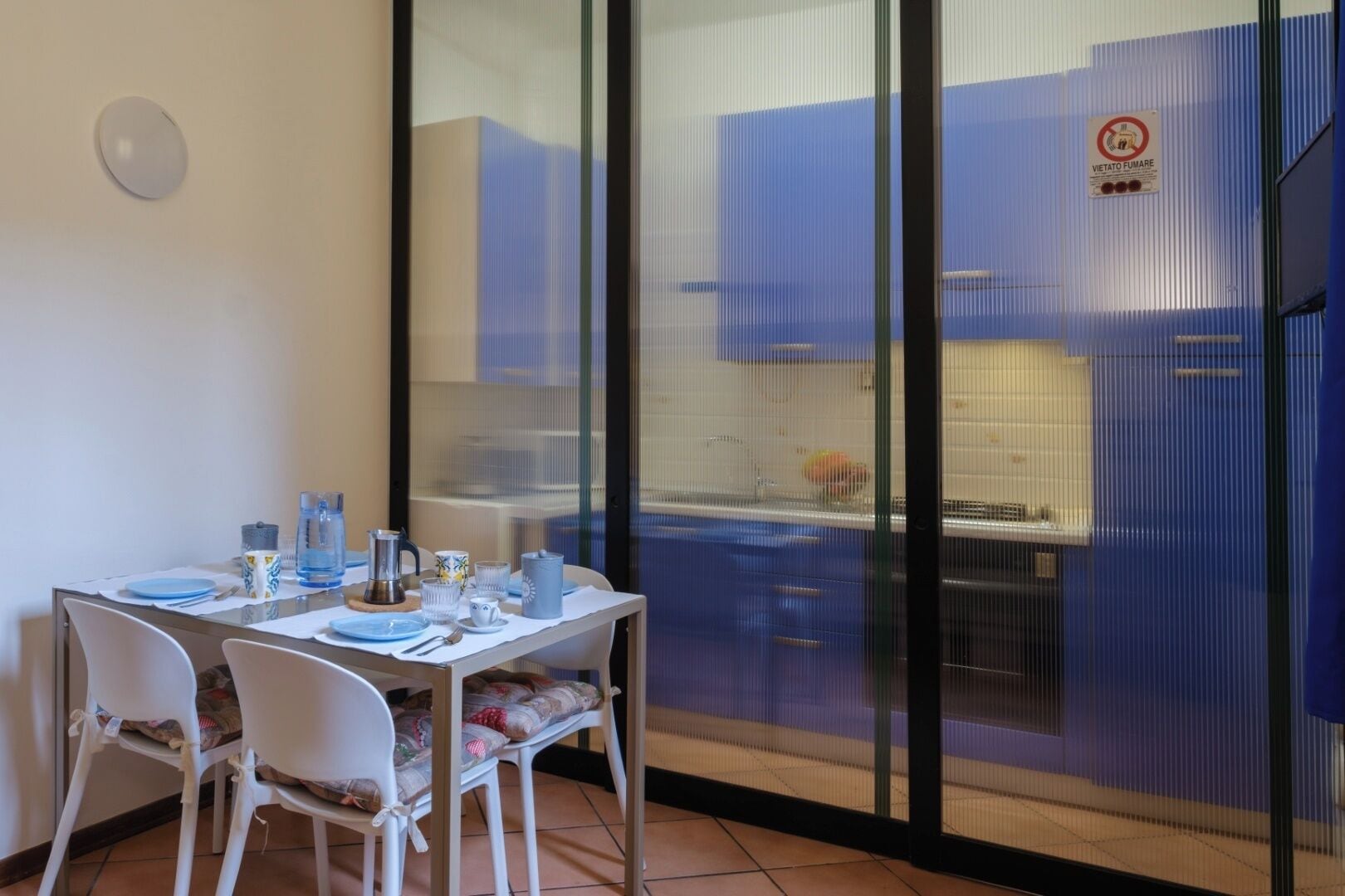 A dining area with a table set for two, chairs, and a view of the kitchen through a glass partition.