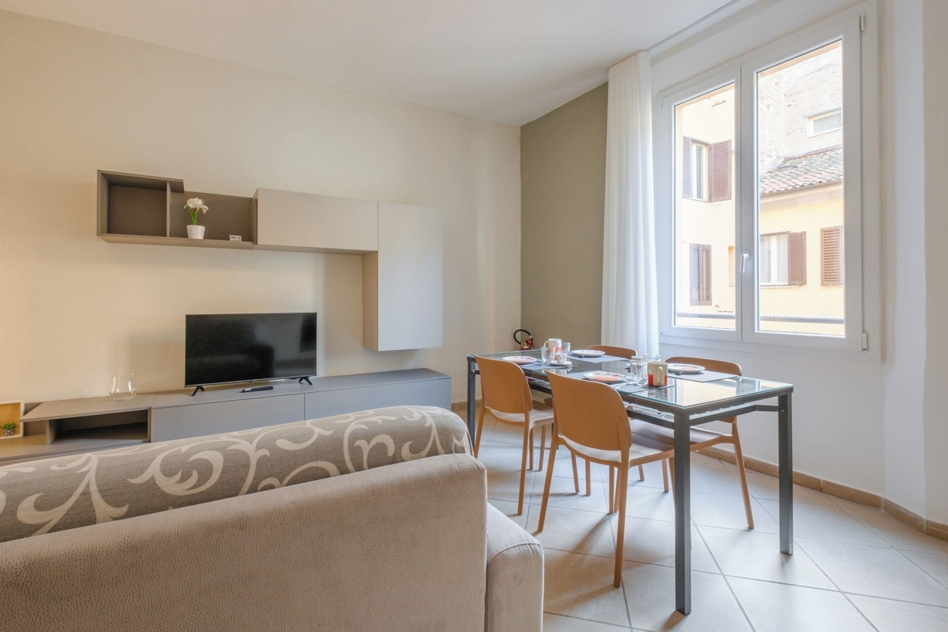 A modern living room with a sofa, a TV, a dining table with chairs, and a window with a view of buildings.