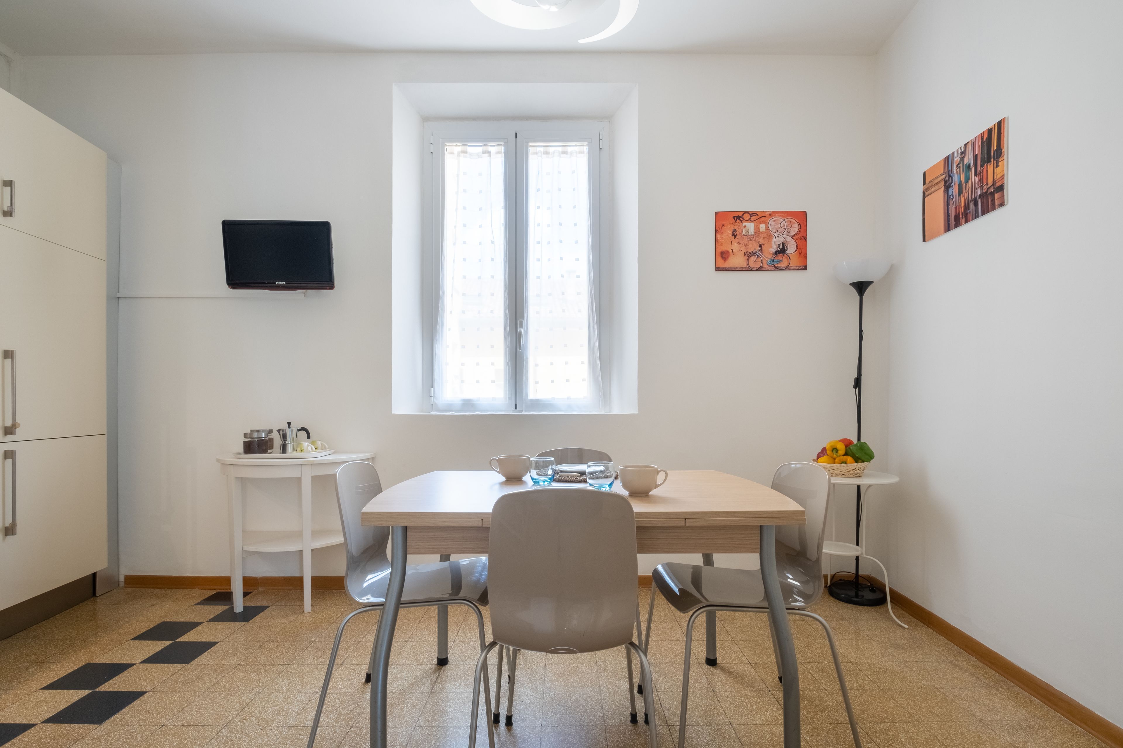 A modern kitchen with white cabinets, a dining table with chairs, a TV mounted on the wall, and a window with curtains.