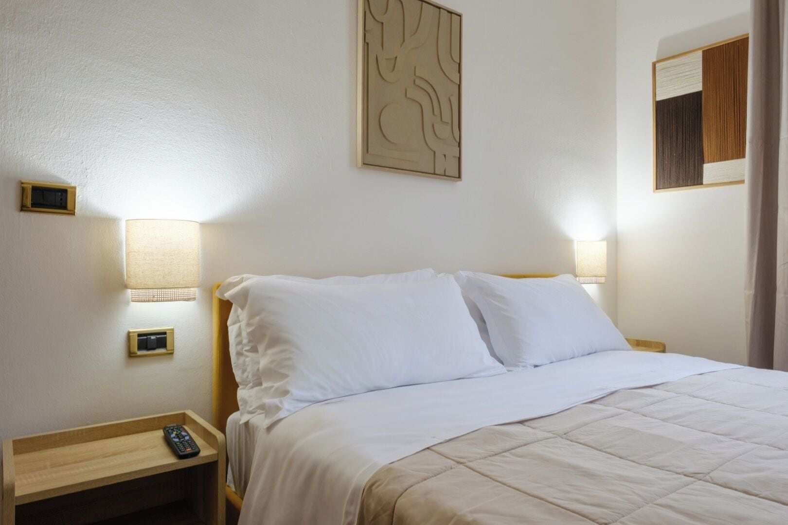 A neatly made bed with white linens, two bedside lamps, and a remote control on a wooden nightstand.