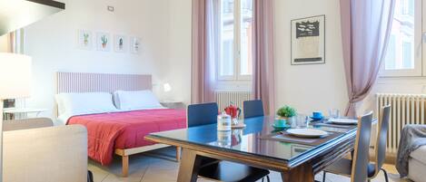 A well-lit room with a bed, a dining table with chairs, a lamp, and a window with curtains.