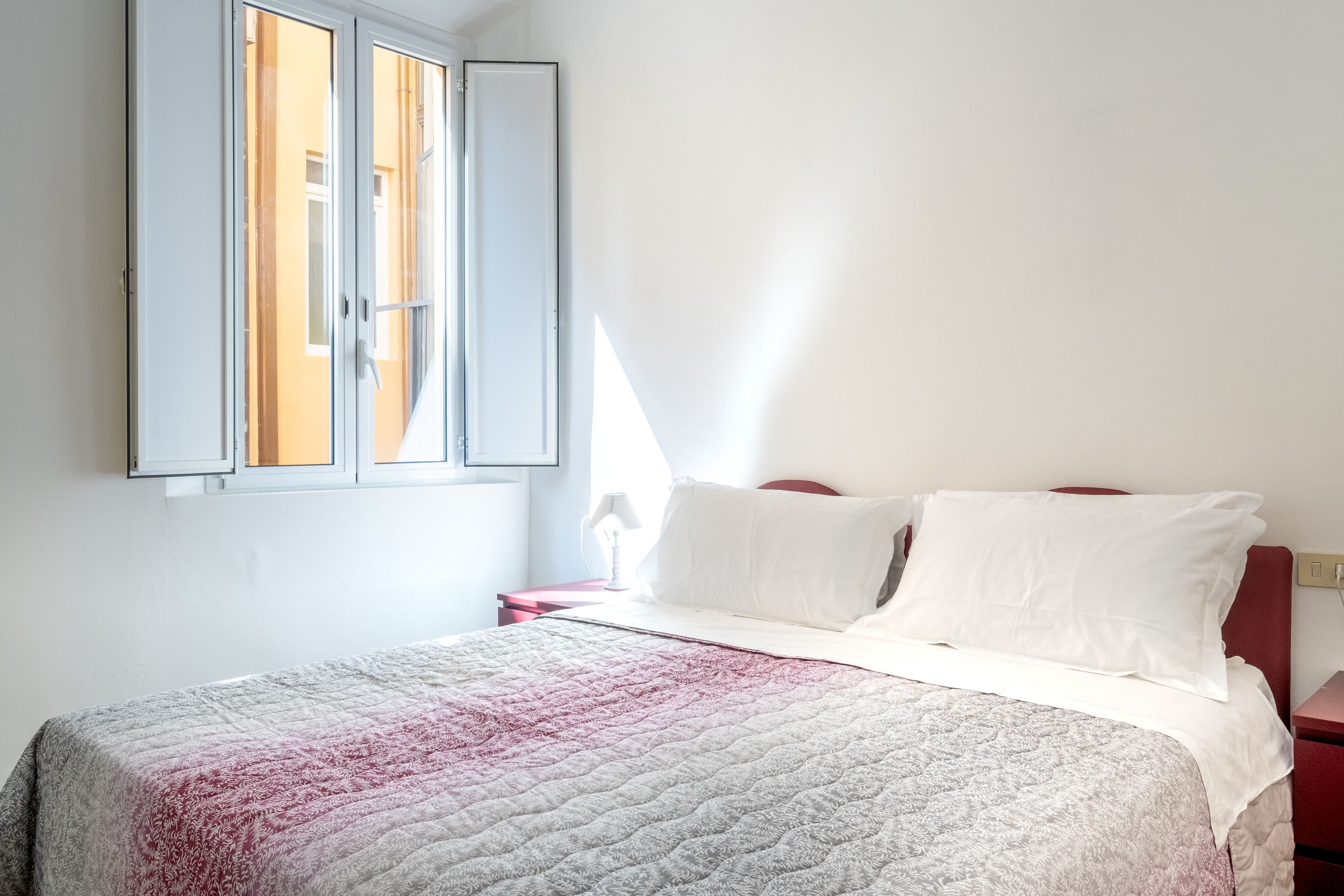 Un dormitorio con una cama, almohadas blancas, una mesita de noche roja y una ventana con cortinas.
