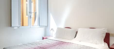 Un dormitorio con una cama, almohadas blancas, una mesita de noche roja y una ventana con cortinas.