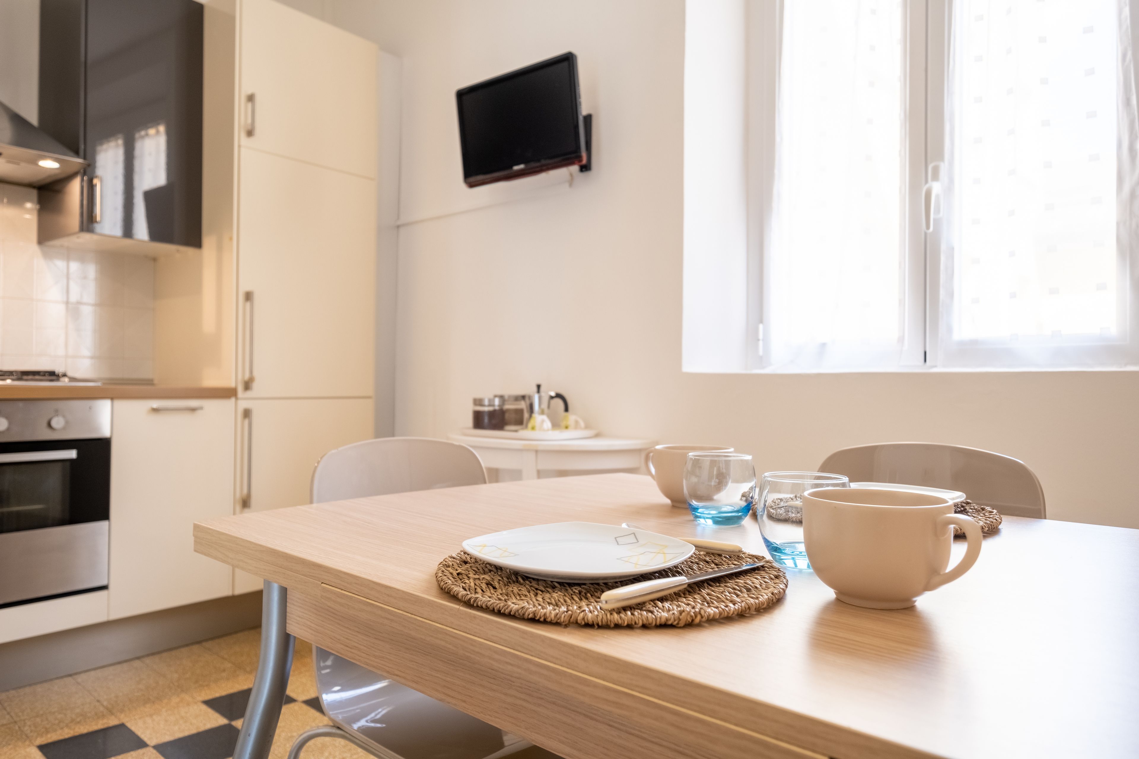 A modern kitchen with a dining area, a TV mounted on the wall, and a window providing natural light.