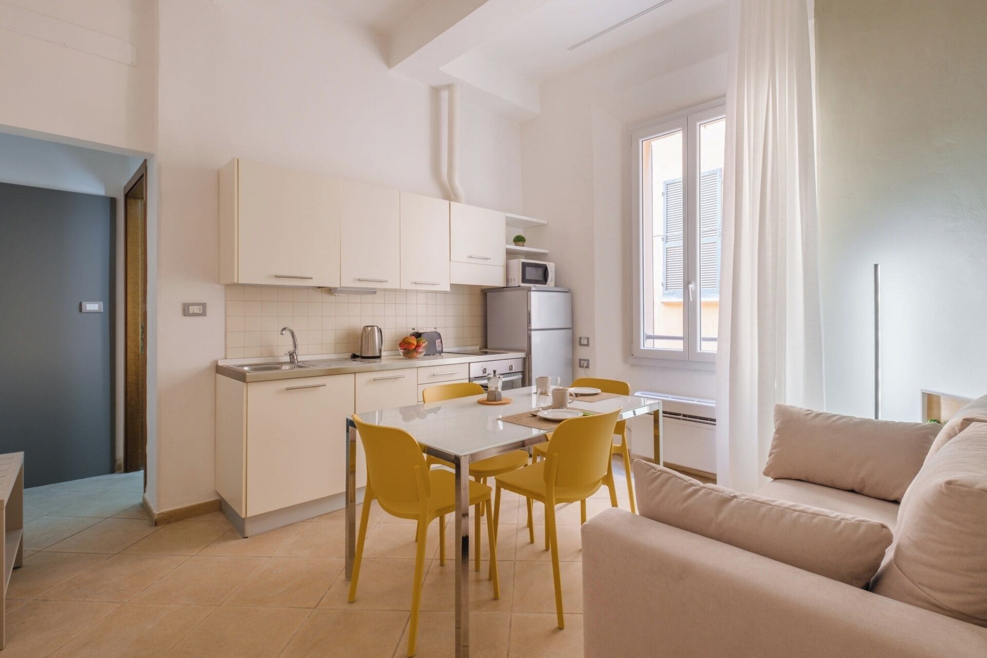 A modern kitchen with white cabinets, a dining table with yellow chairs, and a beige sofa in a bright living area with a large window.