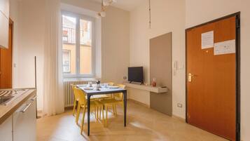 A small, bright kitchen with a dining table and chairs, a TV on a shelf, and a door with a notice.