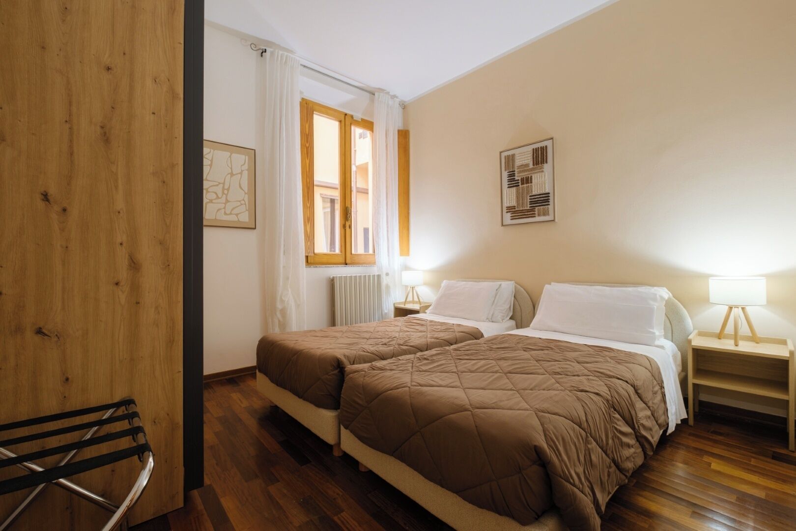 A hotel room with two beds, wooden flooring, a window with curtains, a radiator, and a wall-mounted picture.