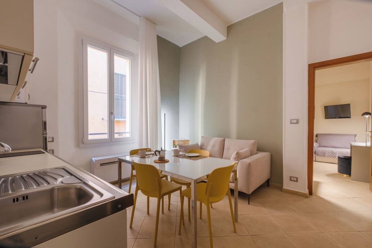 A compact kitchen with a sink, dining table, and chairs. A sofa and a bed are visible in the adjacent living area.
