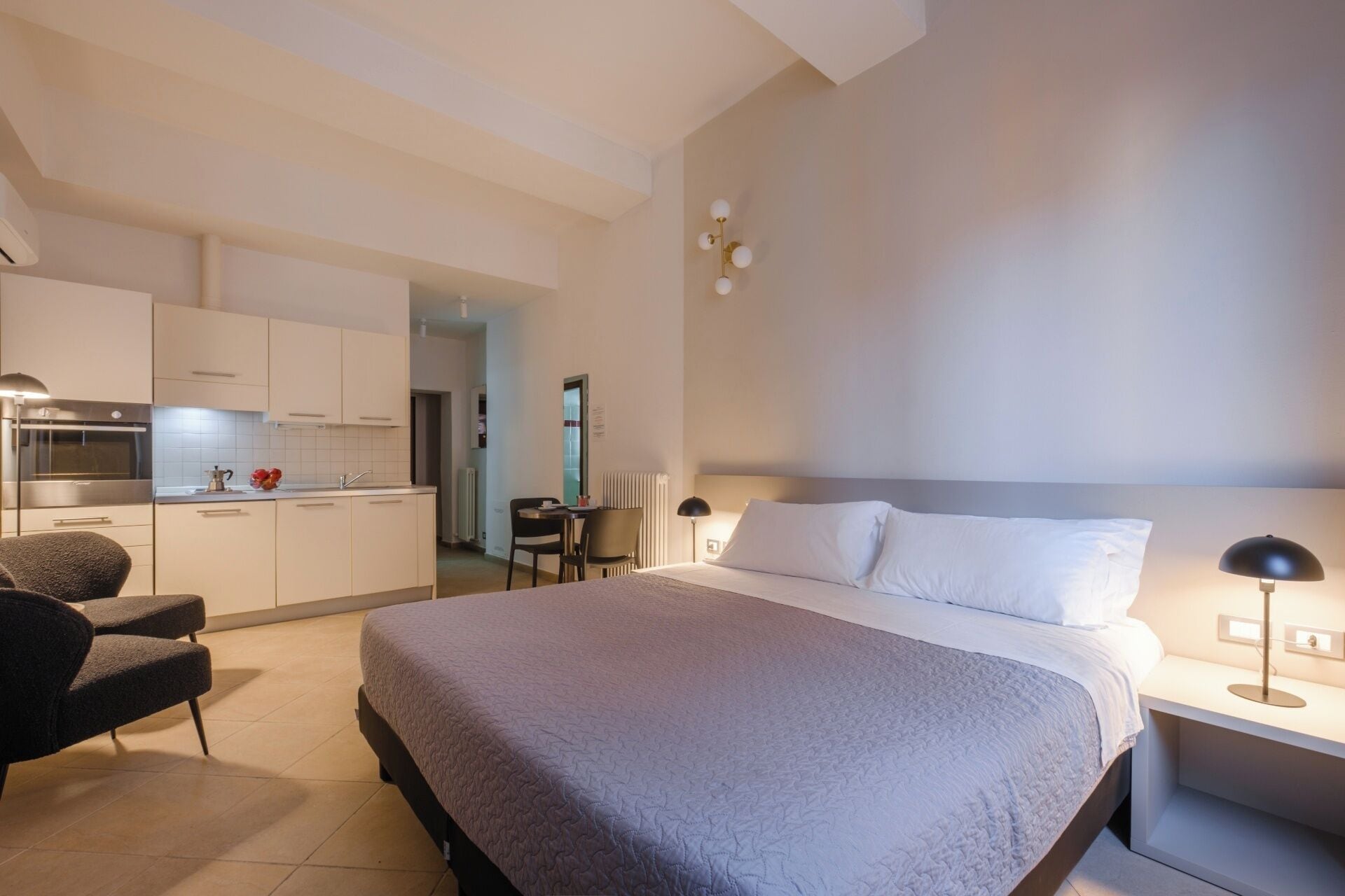 A modern hotel room with a large bed, a desk, a chair, a kitchenette, and a bathroom.
