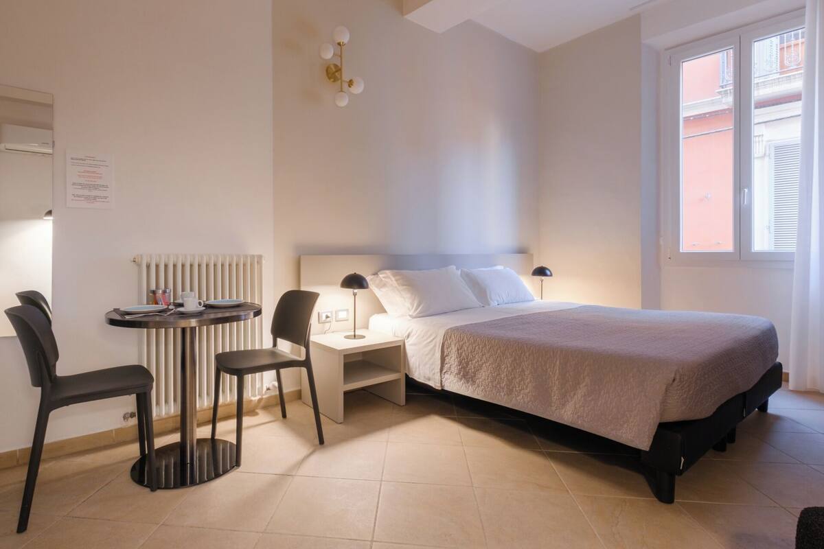 A hotel room with a bed, a desk, a chair, a radiator, and a window with blinds.