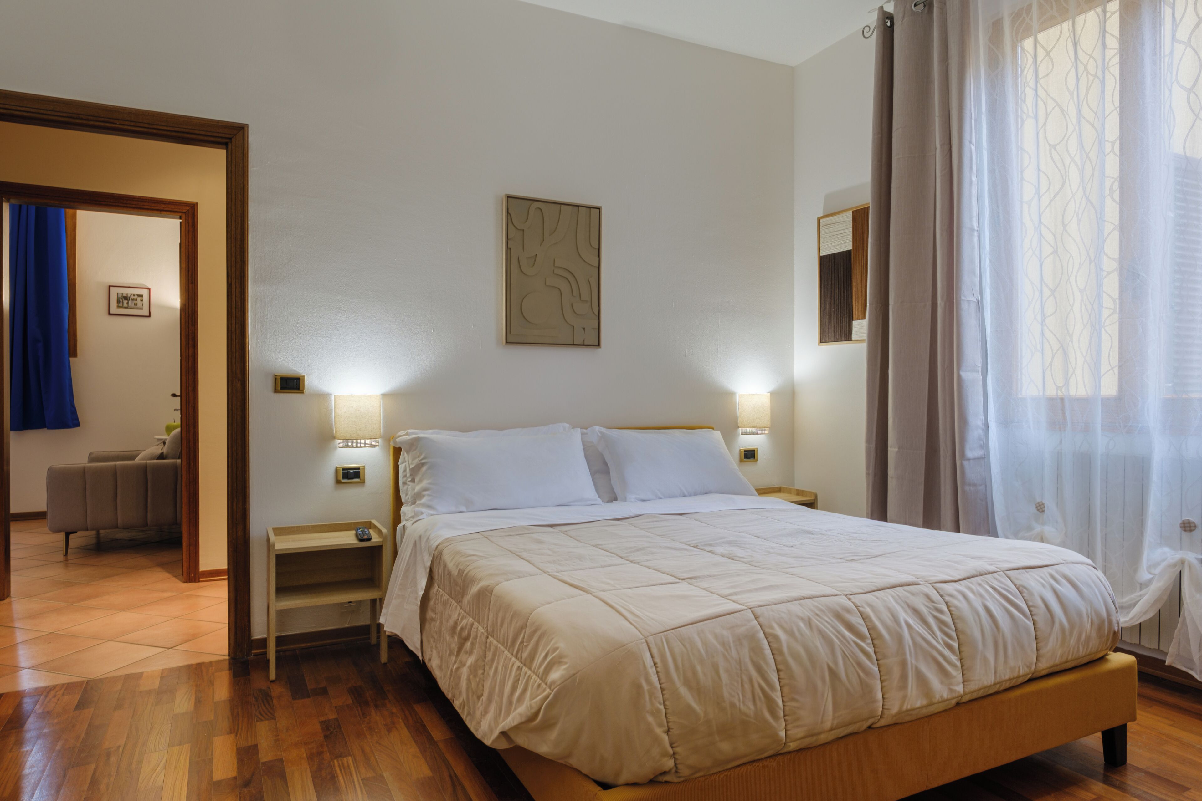 Un dormitorio con cama, mesitas de noche, un cuadro en la pared y vistas a otra habitación con una cortina azul.