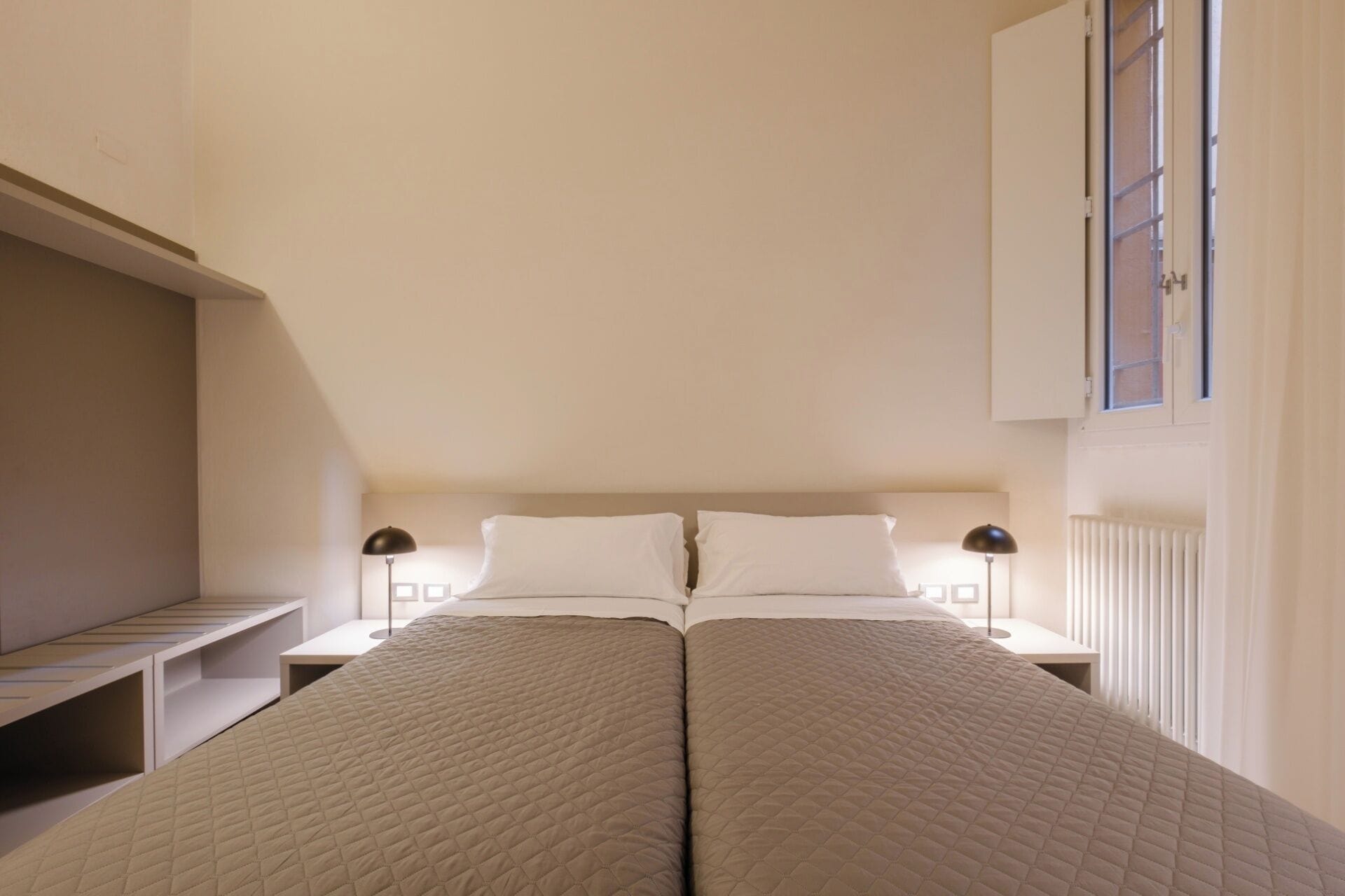 A bedroom with a bed, two bedside tables, a window, and a radiator.