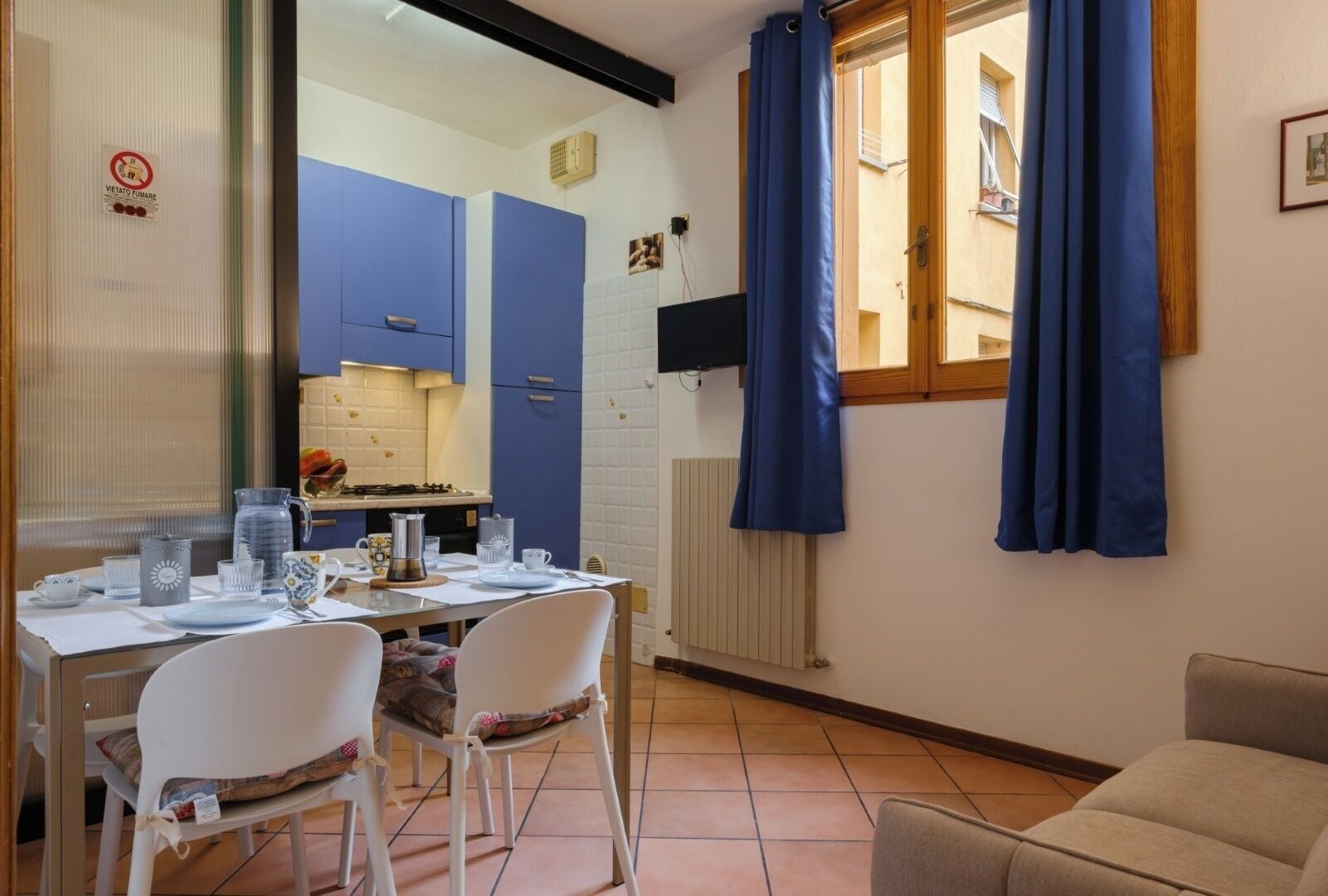 Ein Essbereich mit einem für zwei Personen gedecktem Tisch, einer Küche im Hintergrund und einem Fenster mit blauen Vorhängen.