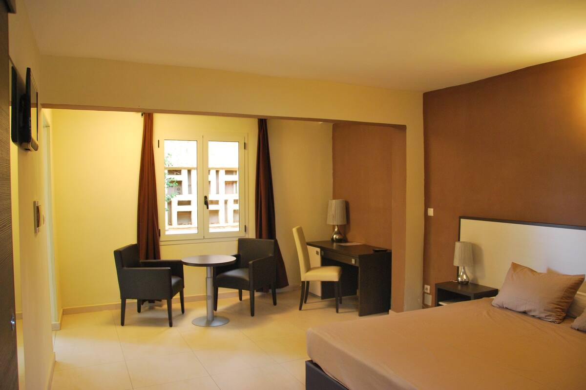 Ein Hotelzimmer mit einem Bett, einem Schreibtisch mit Stuhl, einem kleinen Tisch für zwei Personen, einem an der Wand befestigten Fernseher und einem Fenster mit Vorhängen.