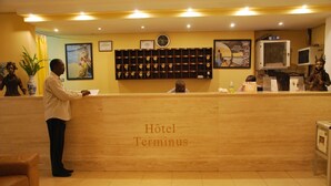Un comptoir d’accueil d’h ôtel avec une horloge, des certificats encadrés et un présentoir mural où sont exposées des photos encadrées.