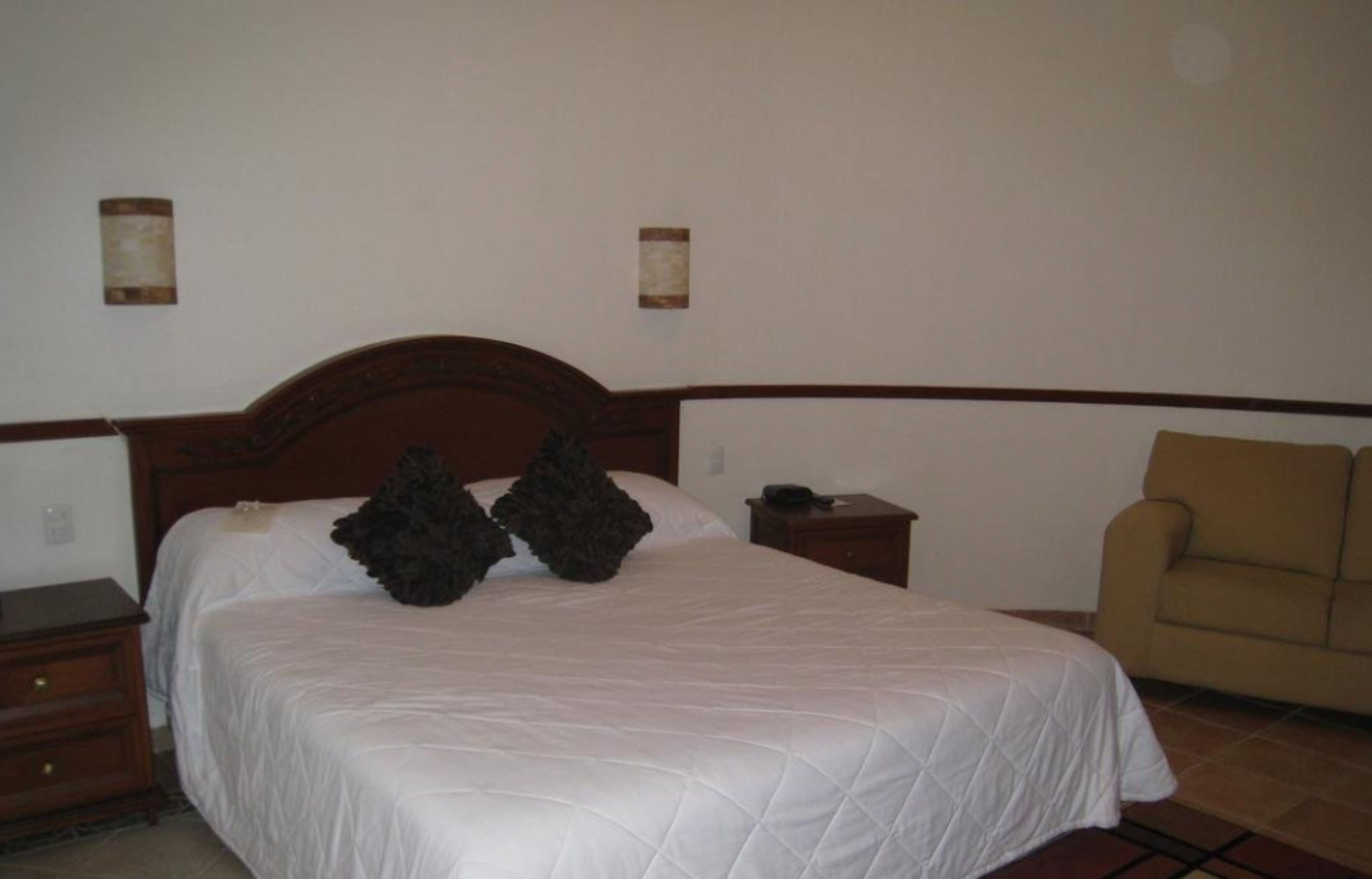 Una cama bien hecha con cabecera de madera, una mesita de noche, una silla y dos lámparas fijadas a la pared.