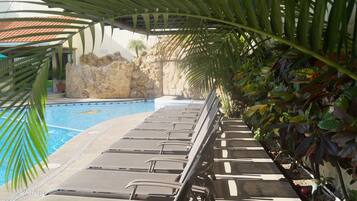 Un área junto a la piscina con sillas de descanso, un muro de piedra y plantas tropicales.