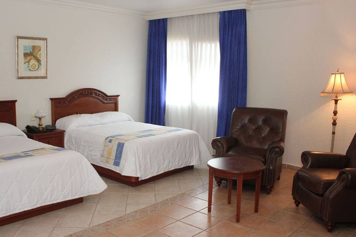 Quarto de hotel com duas camas, uma cadeira, uma mesinha e um abajur.