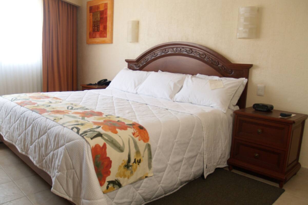 Cama bem arrumada com colcha floral, cabeceira de madeira, mesa de cabeceira com telefone e mesa de apoio com abajur.