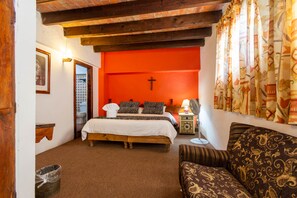 Une chambre à coucher avec un lit, un banc en bois, un canapé, un mur orné d’une croix et une fenêtre avec des rideaux.