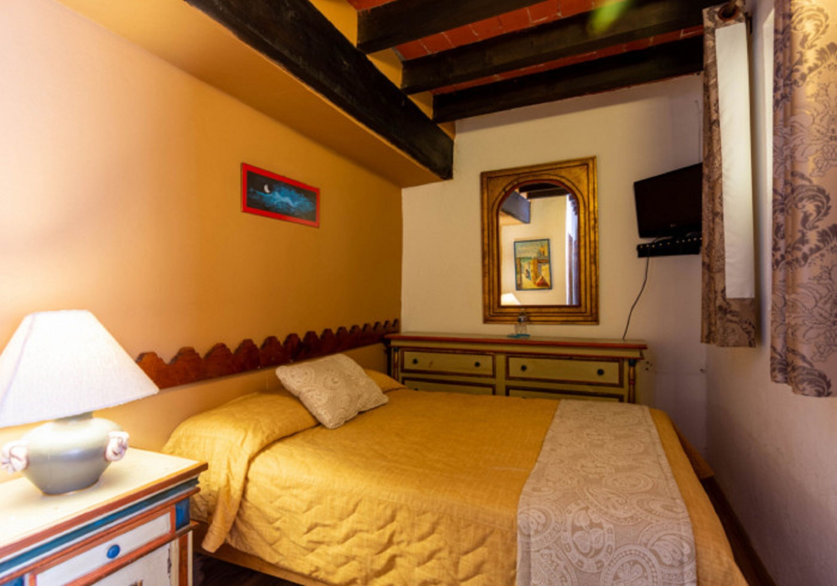 Une chambre à coucher avec un lit, une table de chevet, une lampe, une commode, un miroir et un téléviseur fixé au mur.