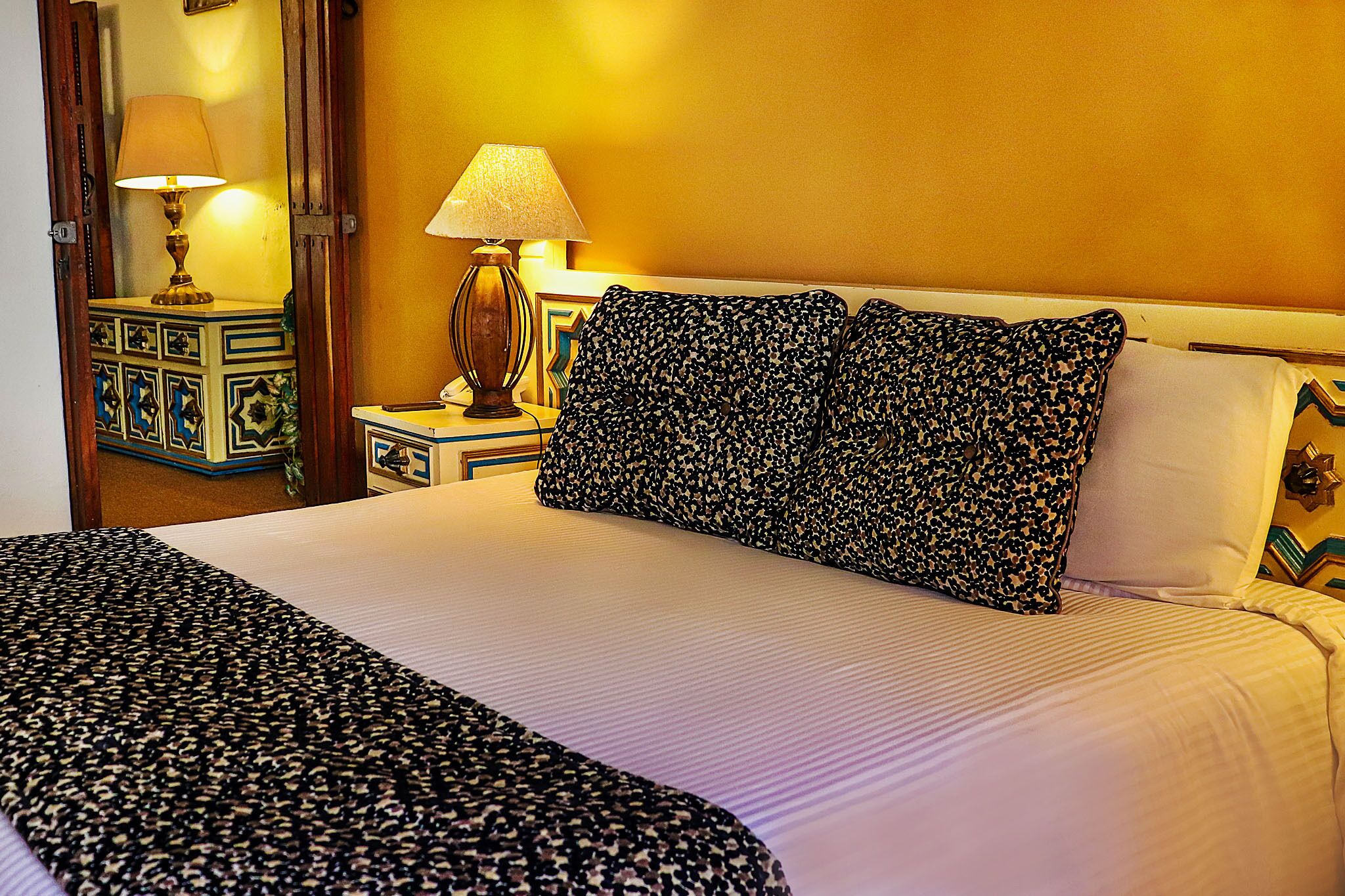 A hotel room with a large bed, two pillows with a leopard print, a bedside lamp, and a decorative nightstand.
