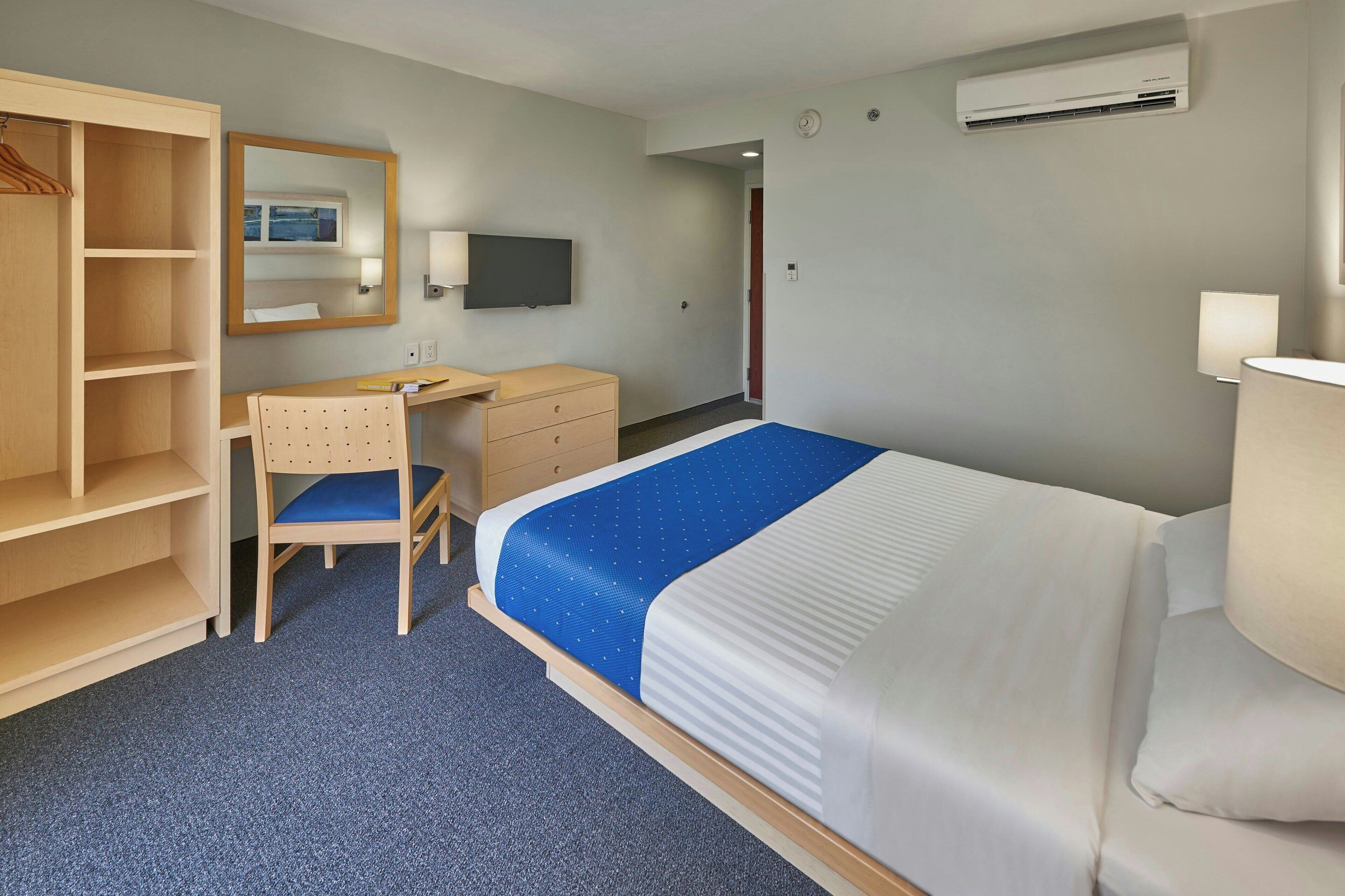 Una habitación de hotel con una cama, un escritorio, una silla, un armario, un espejo, un televisor y un equipo de aire acondicionado.
