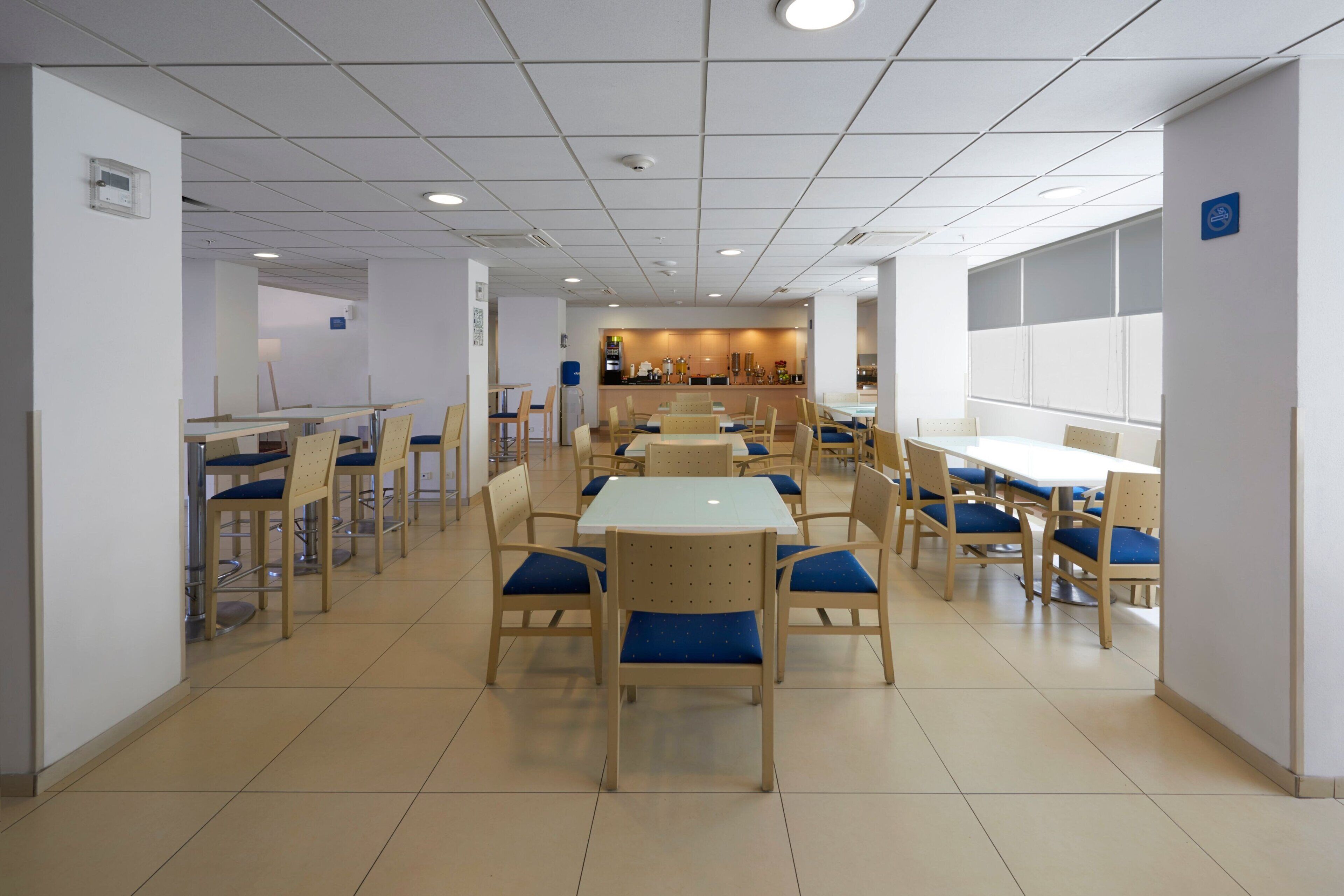 A spacious cafeteria with multiple tables and chairs, a central serving area, and a clean, tiled floor.