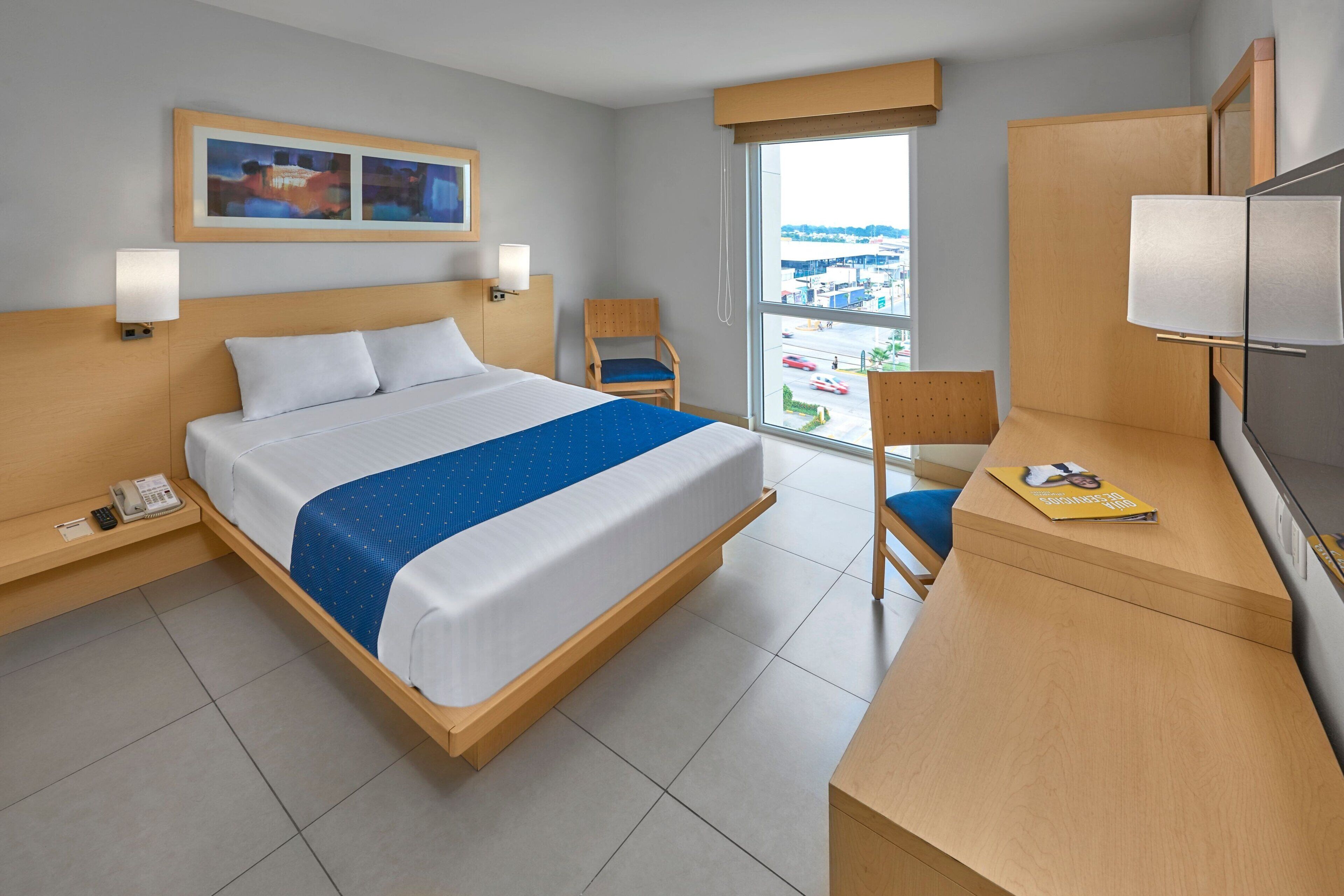 A hotel room with a bed, bedside tables, a desk, a chair, a TV, and a window with a view of buildings.