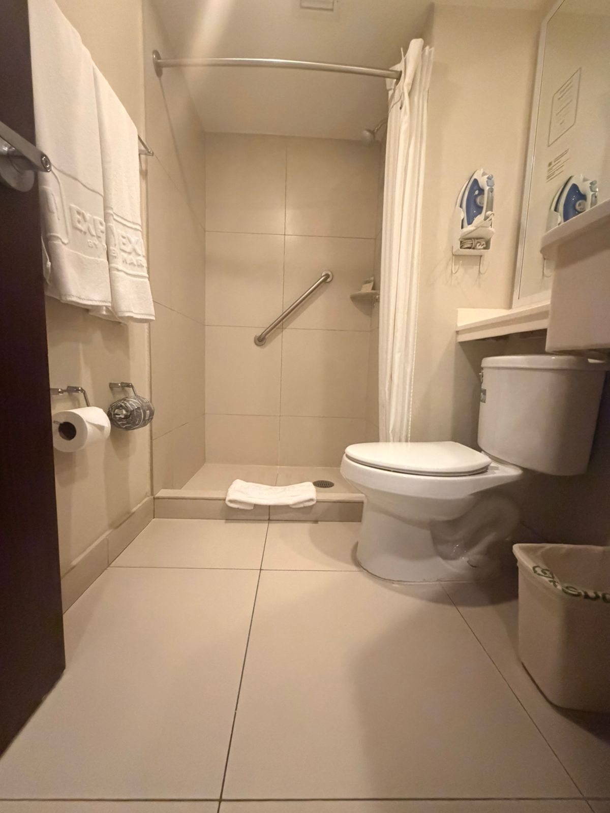 Un baño con inodoro, ducha con barra de apoyo, toallero y cubo de basura.