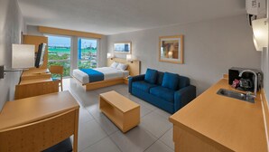 A modern hotel room with a bed, a blue sofa, a wooden desk, a small kitchen area, and a large window offering a view of the outdoors. - City Express by Marriott Xalapa (Xalapa)