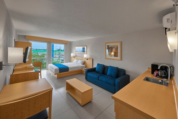 A modern hotel room with a bed, a blue sofa, a wooden desk, a small kitchen area, and a large window offering a view of the outdoors. - City Express by Marriott Xalapa (Xalapa)