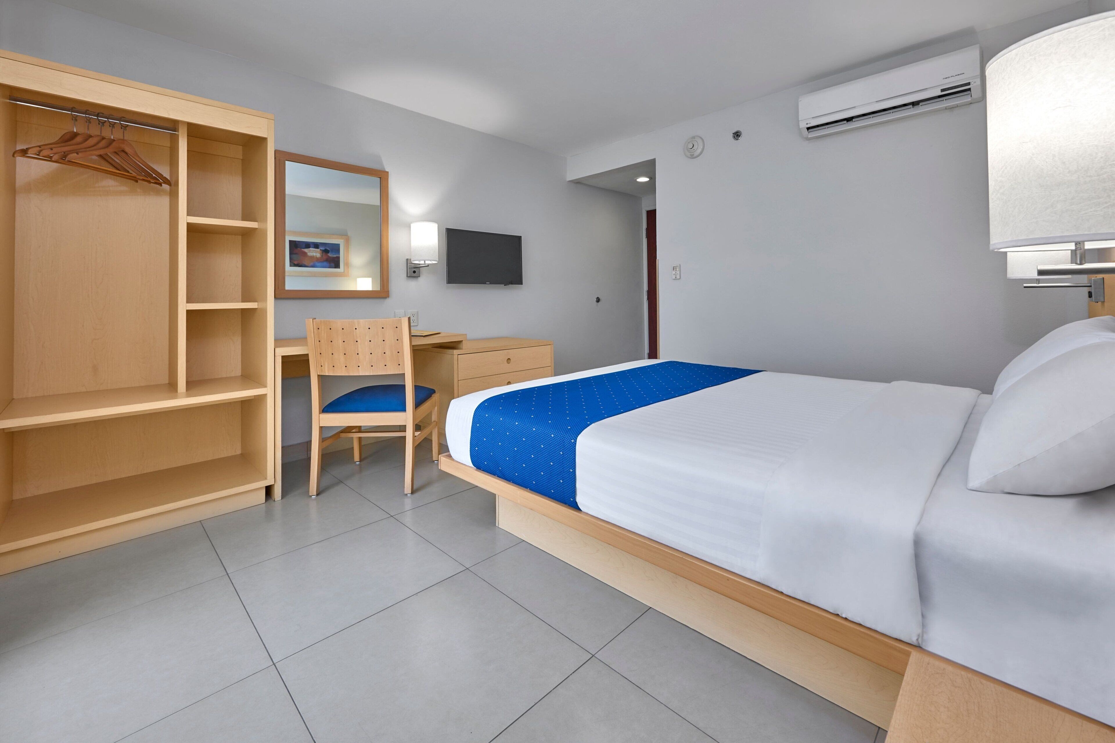 A hotel room with a bed, a wooden wardrobe, a desk, a chair, a television, and an air conditioning unit.