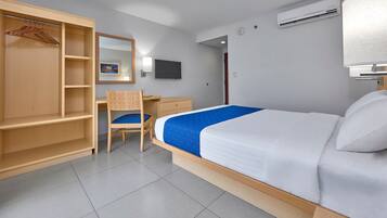 A hotel room with a bed, a wooden wardrobe, a desk, a chair, a television, and an air conditioning unit.