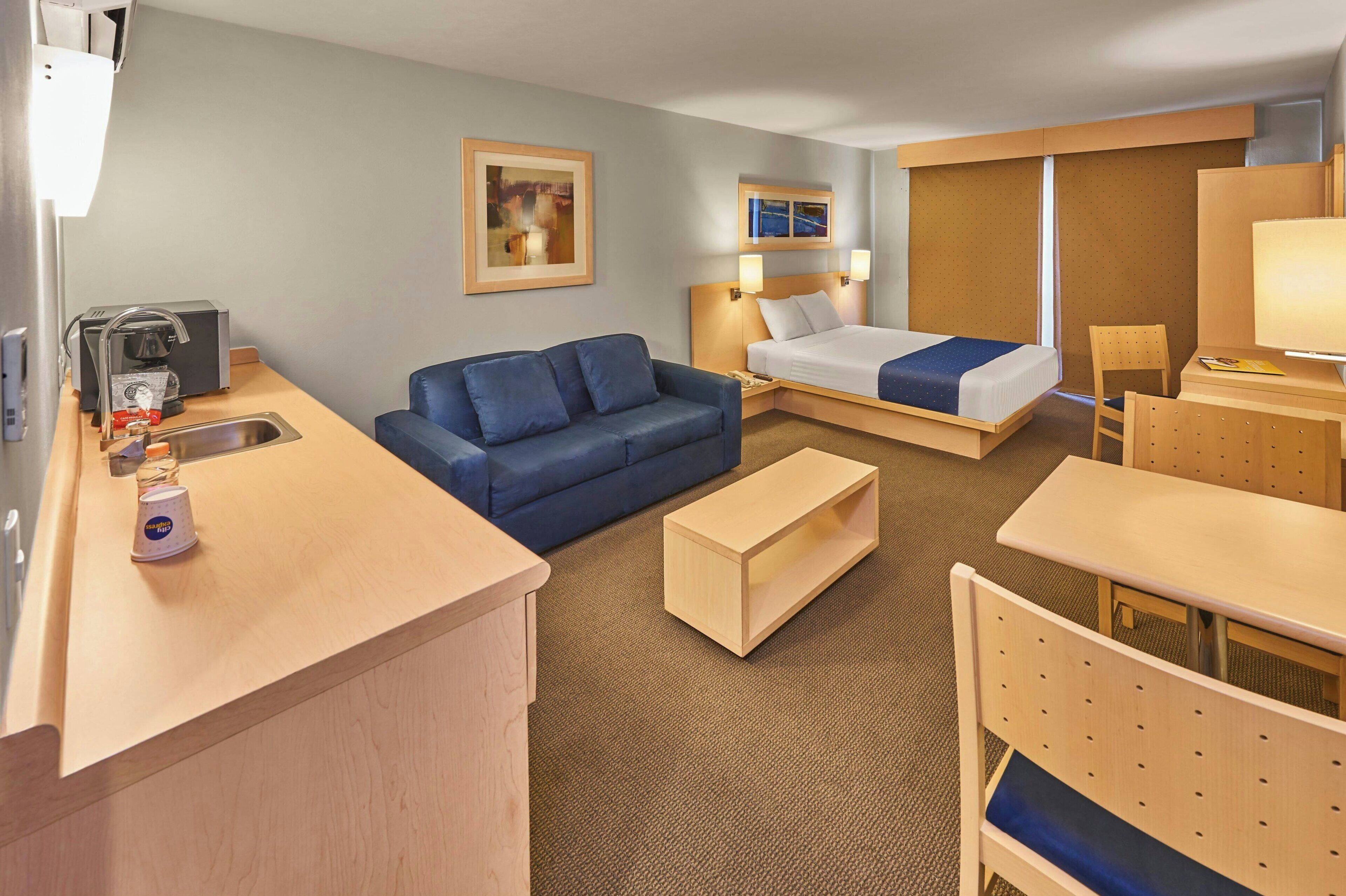 A hotel room with a bed, a sofa, a desk, a chair, a coffee machine, and a sink.