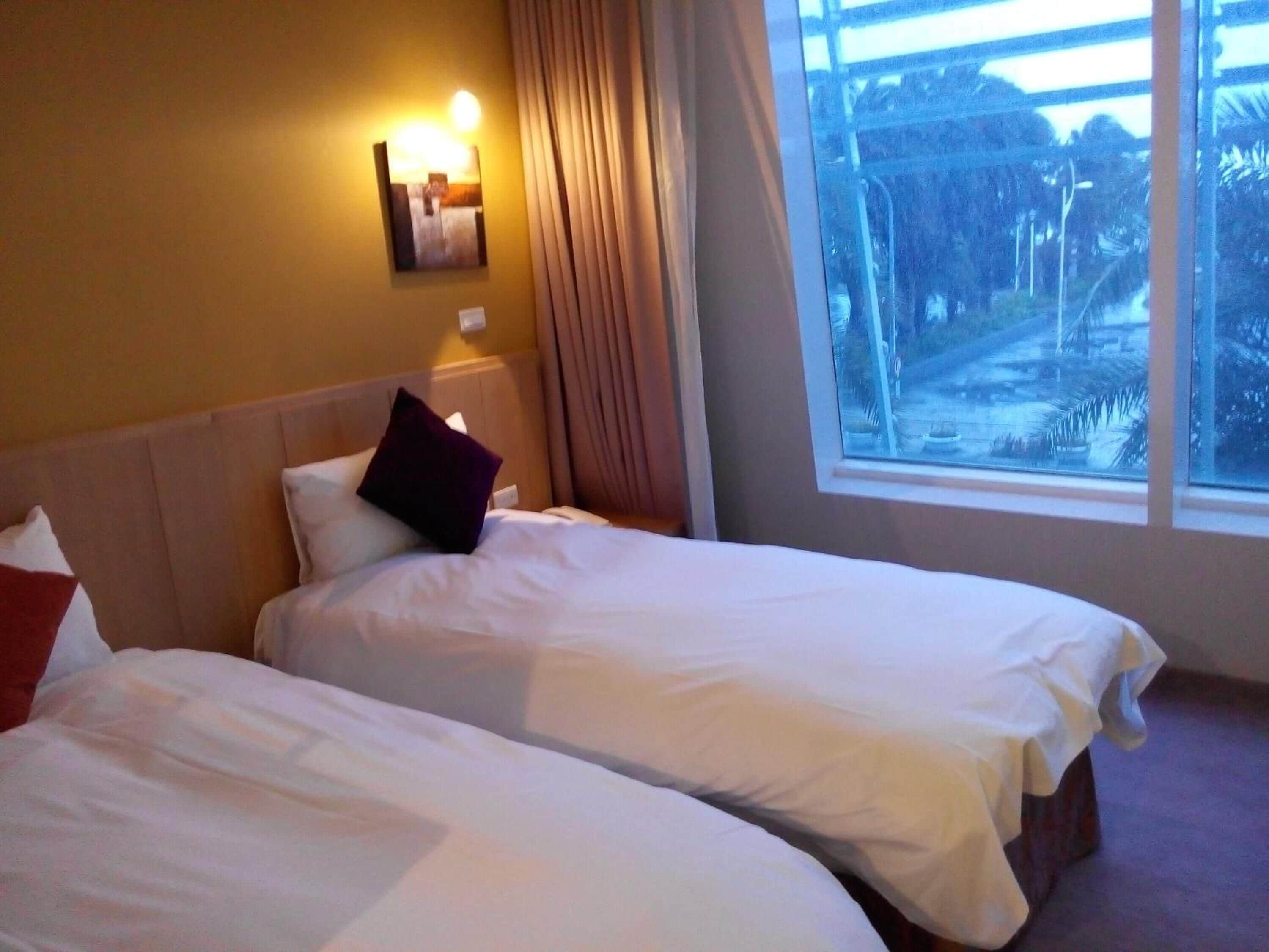 Een hotelkamer met twee bedden, een nachtkastje, een wandlamp en een raam met uitzicht op groen.