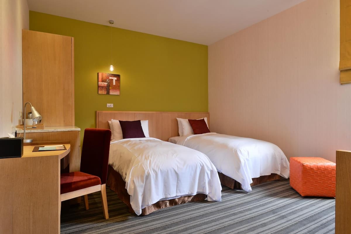 Habitación de hotel con dos camas, un escritorio con una silla, un reposapiés naranja pequeño y un cuadro en la pared.