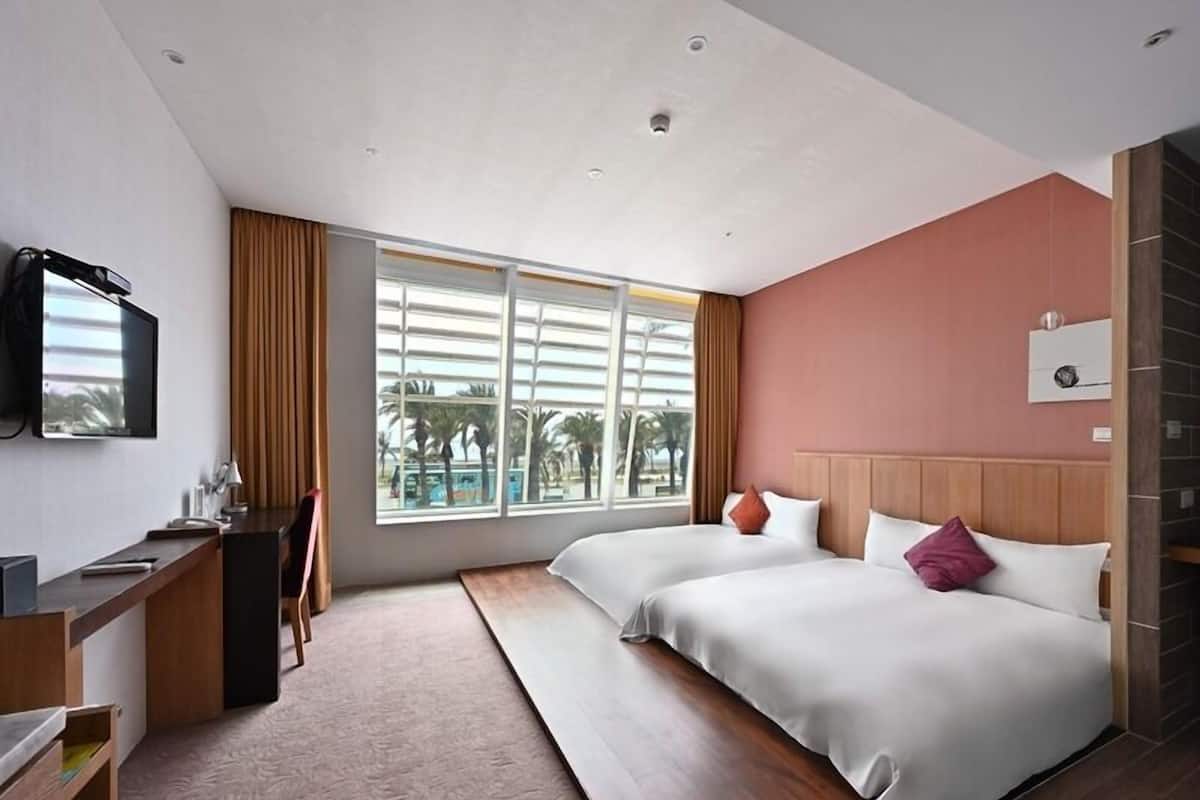 Una habitación de hotel con cama, escritorio, televisor y ventana con vista a palmeras.
