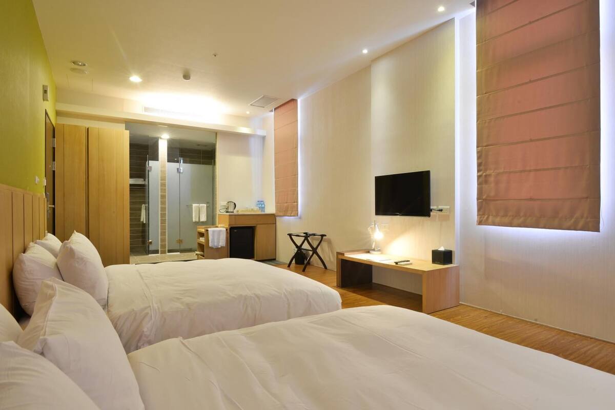 Habitación de hotel con dos camas, un escritorio, un televisor y un baño visible a través de una puerta abierta.