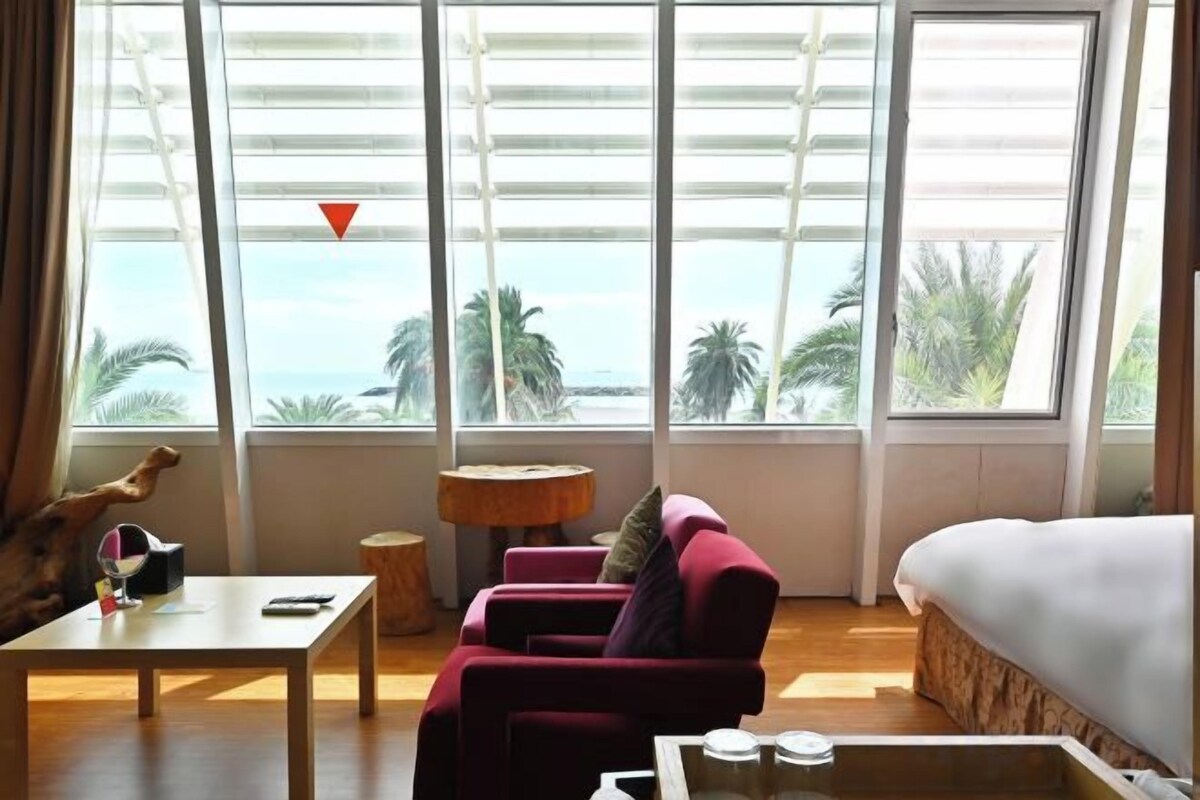 A room with a bed, a red armchair, a wooden table, and a view of the ocean through large windows.