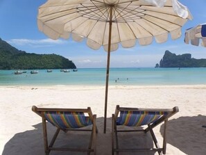 Deux chaises de plage en bois, dotées de coussins rayés aux couleurs vives, sont placées sous un grand parasol sur une plage de sable fin. L’eau est d’un bleu turquoise limpide et, au loin, on aperçoit des bateaux.