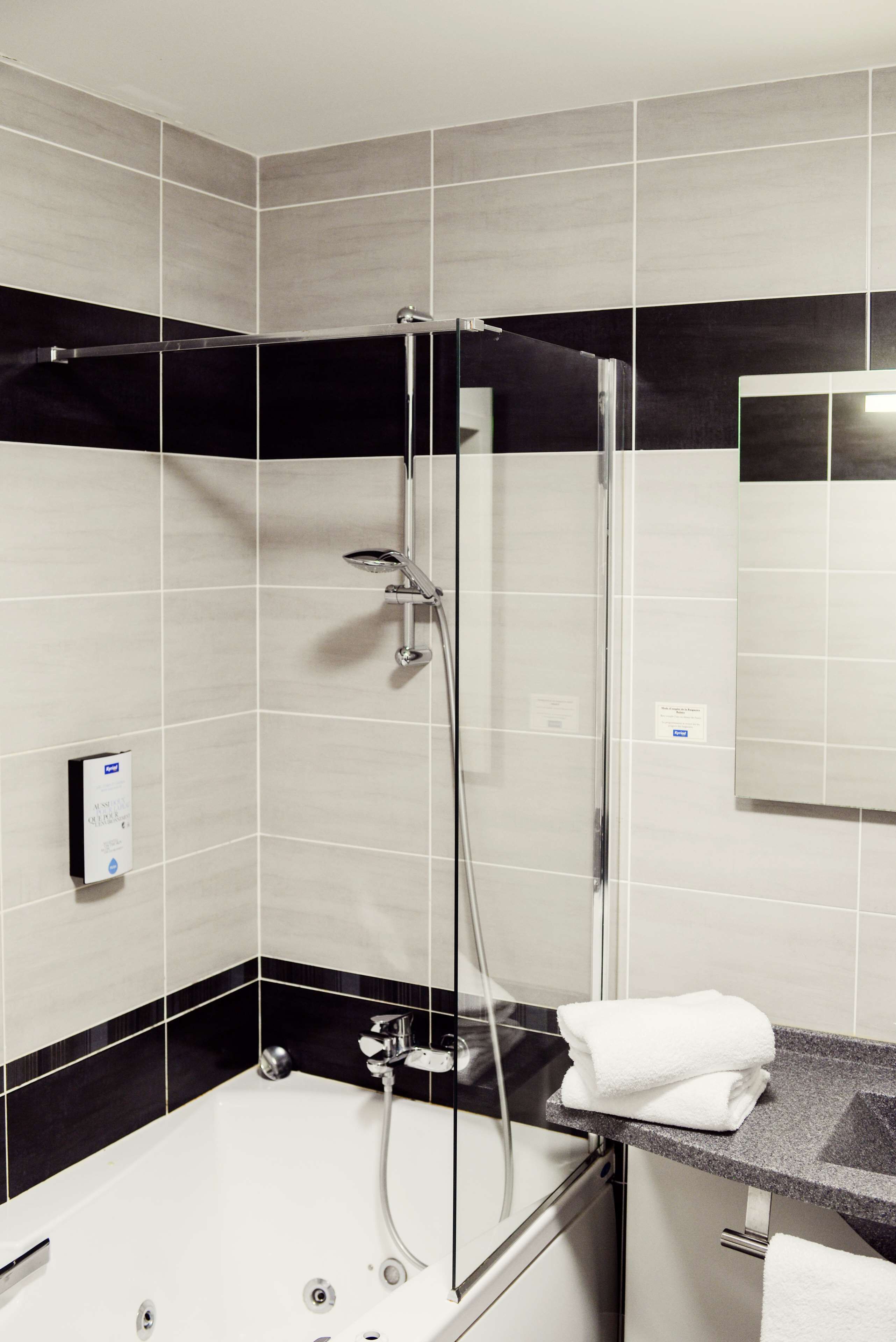 Un baño moderno con ducha de vidrio, pared de azulejos blanco y negro y un lavabo blanco con una toalla sobre la encimera.