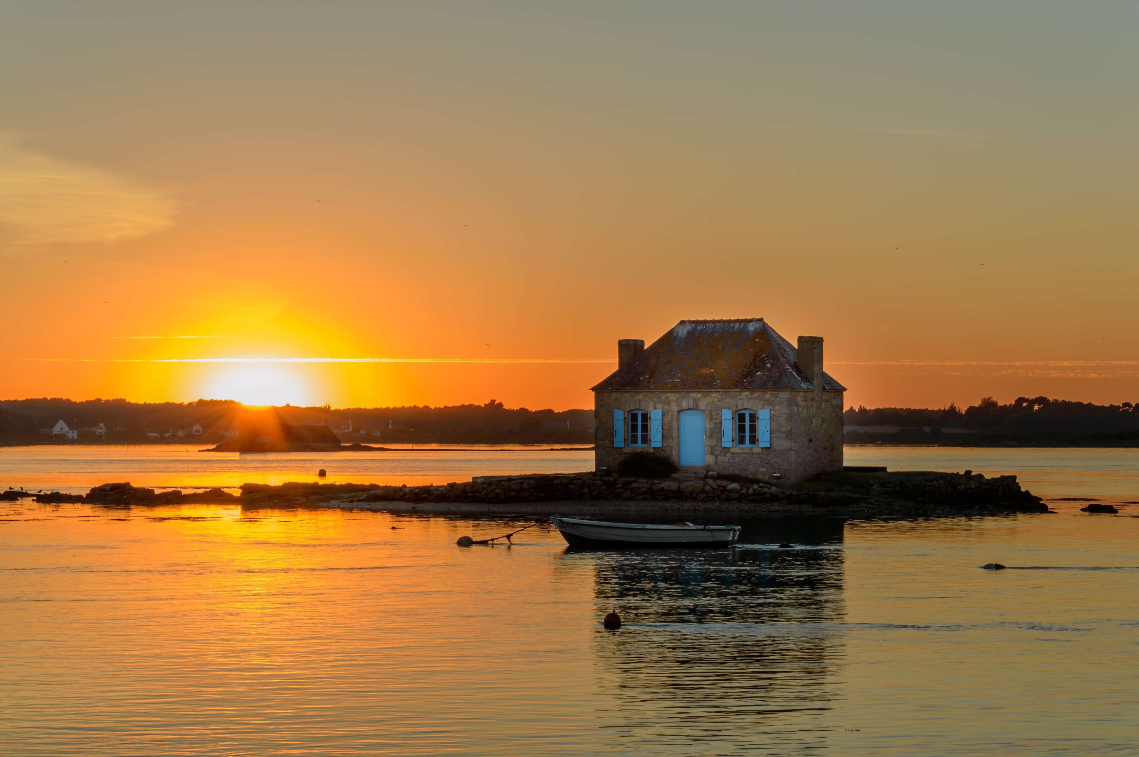 Una casa de piedra con chimenea, ubicada en una pequeña isla en aguas tranquilas al atardecer.