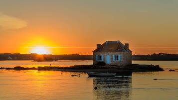 Una casa de piedra con chimenea, ubicada en una pequeña isla en aguas tranquilas al atardecer.