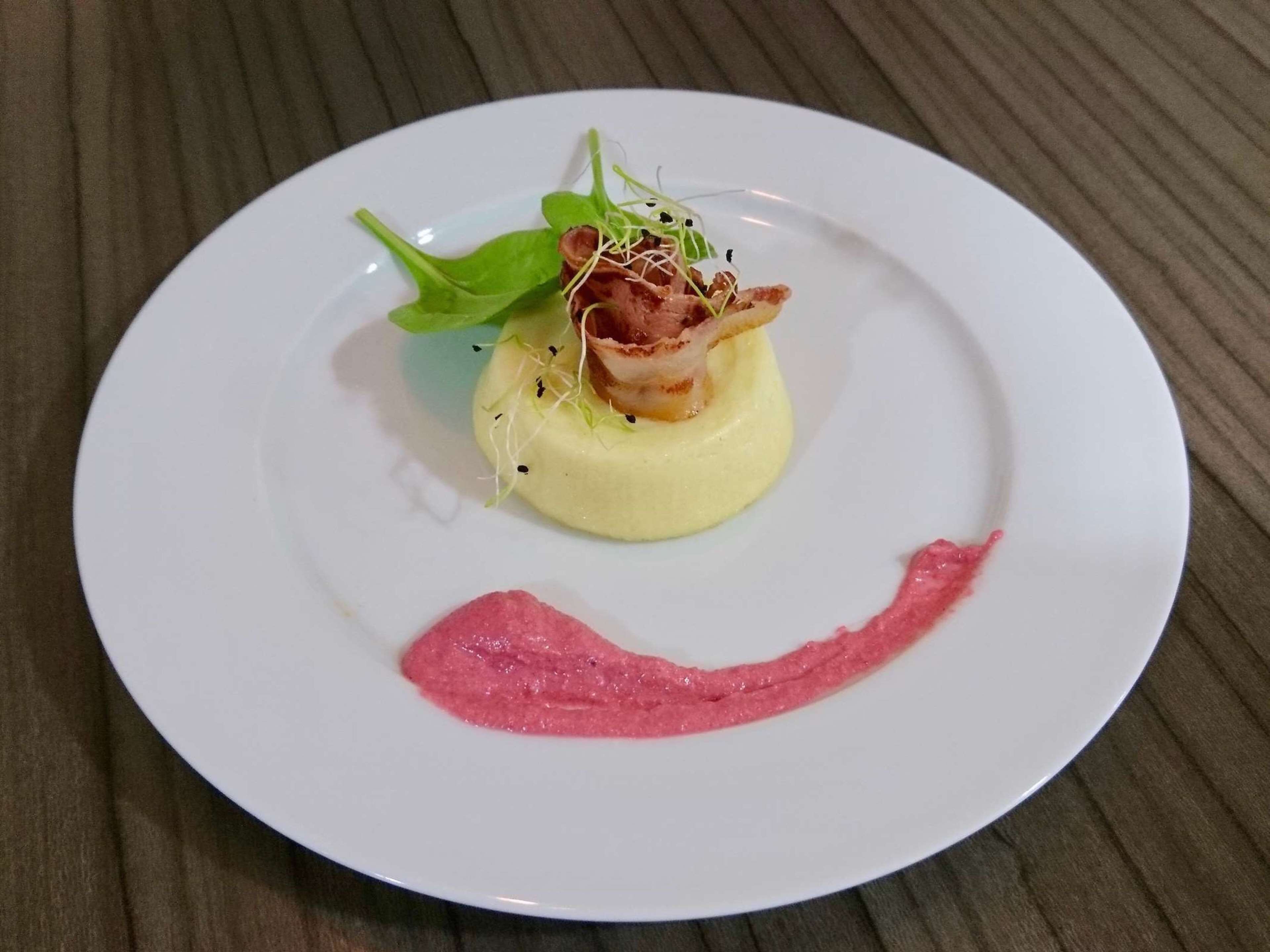 A white plate with a dish consisting of a yellow base, a pink sauce, and a piece of meat with greens on top.