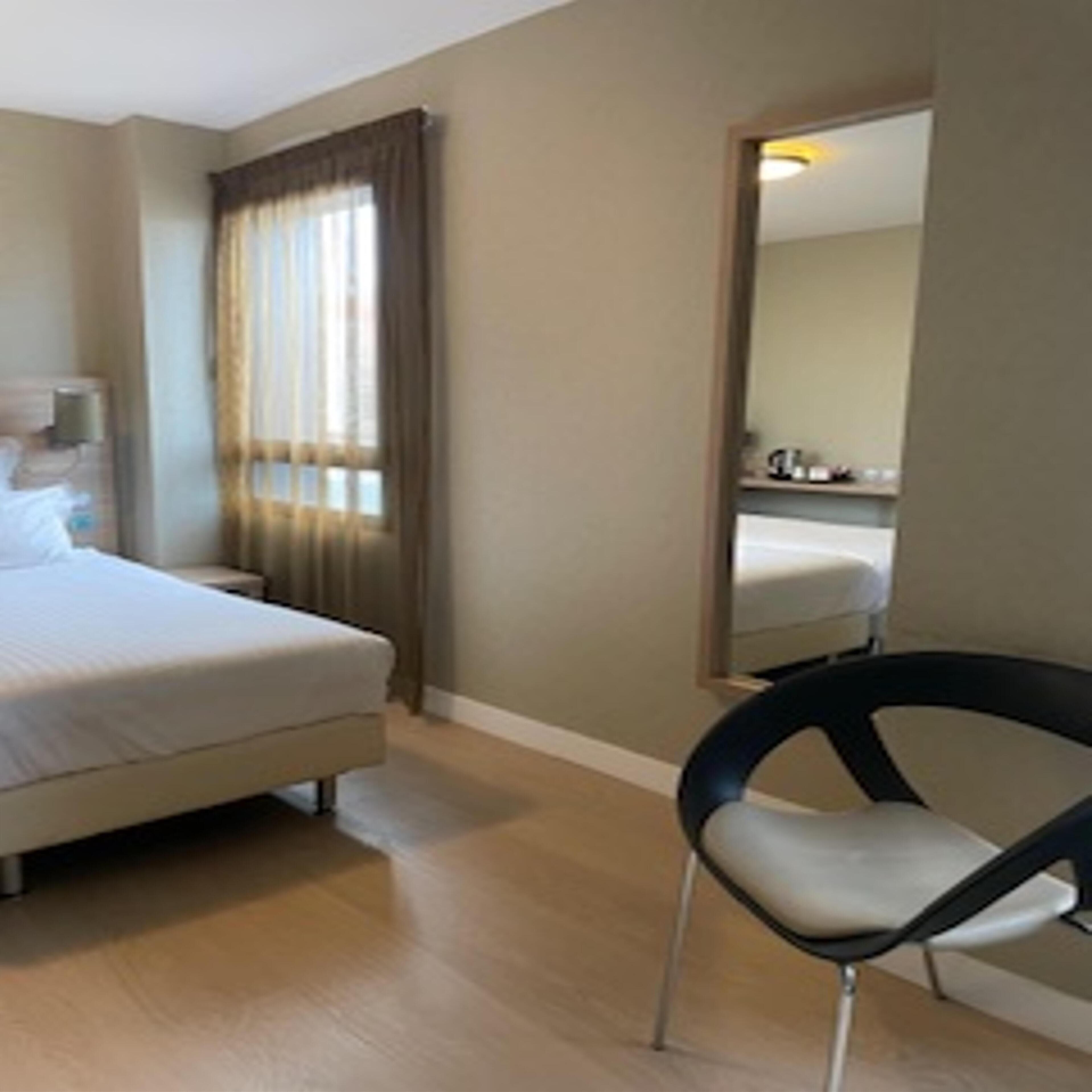 A hotel room with a bed, a chair, a nightstand, a lamp, and a window with curtains.