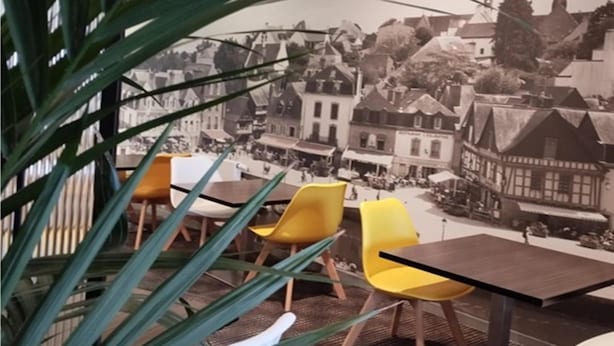 Un café con sillas amarillas y una mesa, con vistas a un pueblo histórico de edificios tradicionales y una aguja de iglesia.