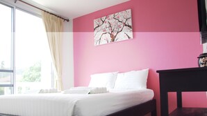 Un dormitorio con cama, mesita de noche, ventana con cortinas y pared rosa con un cuadro floral.