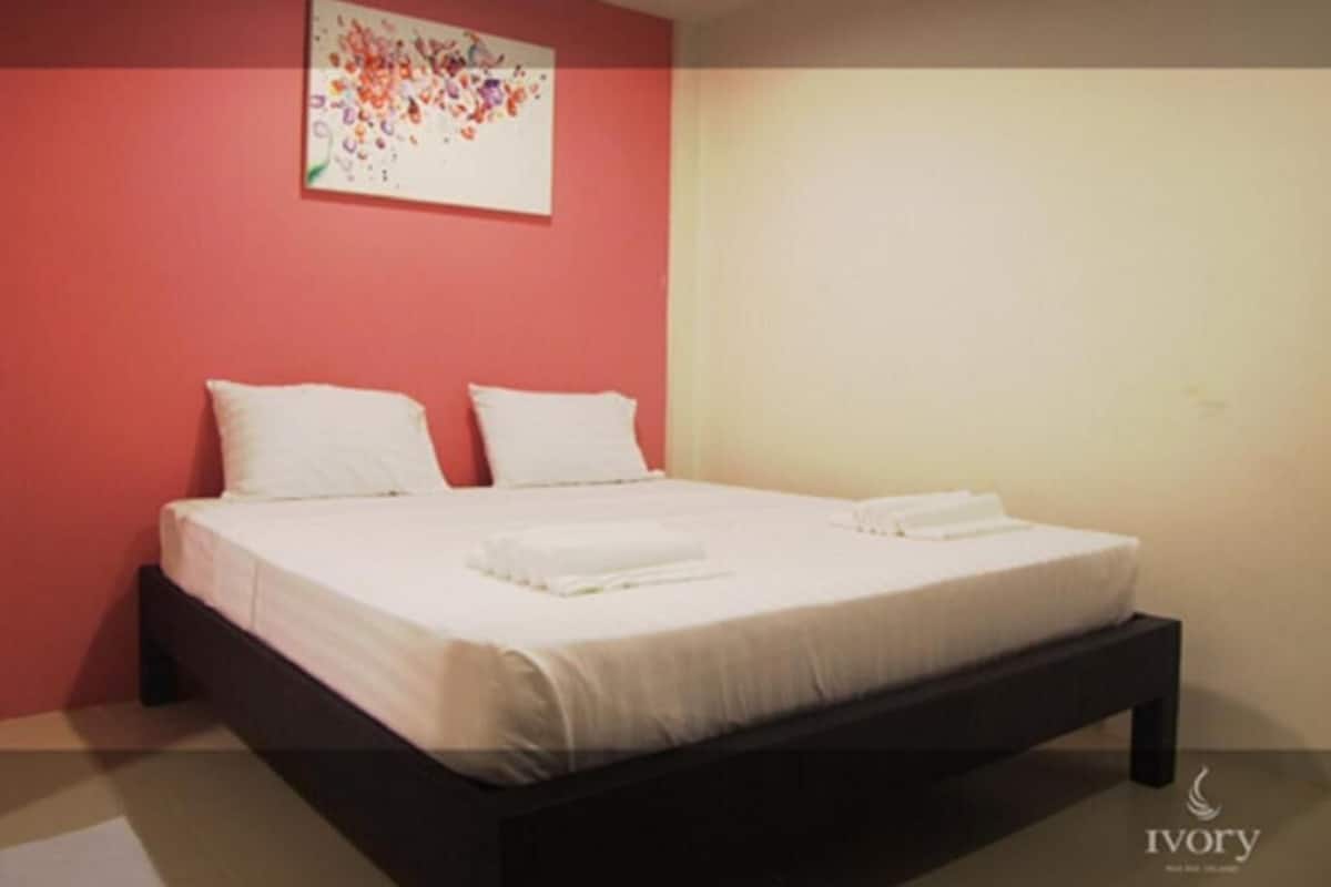 A single bed with white linens and pillows, a dark wooden frame, and a red wall with a floral artwork.