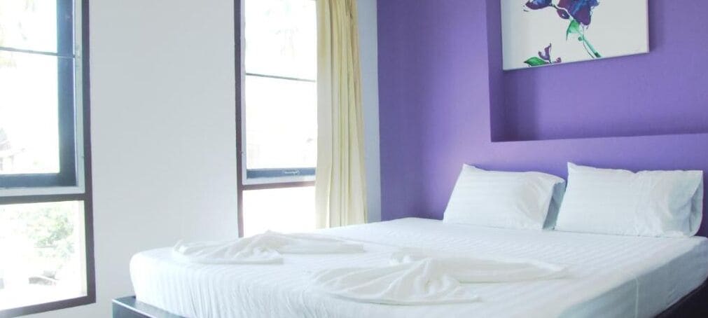 A neatly made bed with white linens in a room with purple walls and a window with curtains.